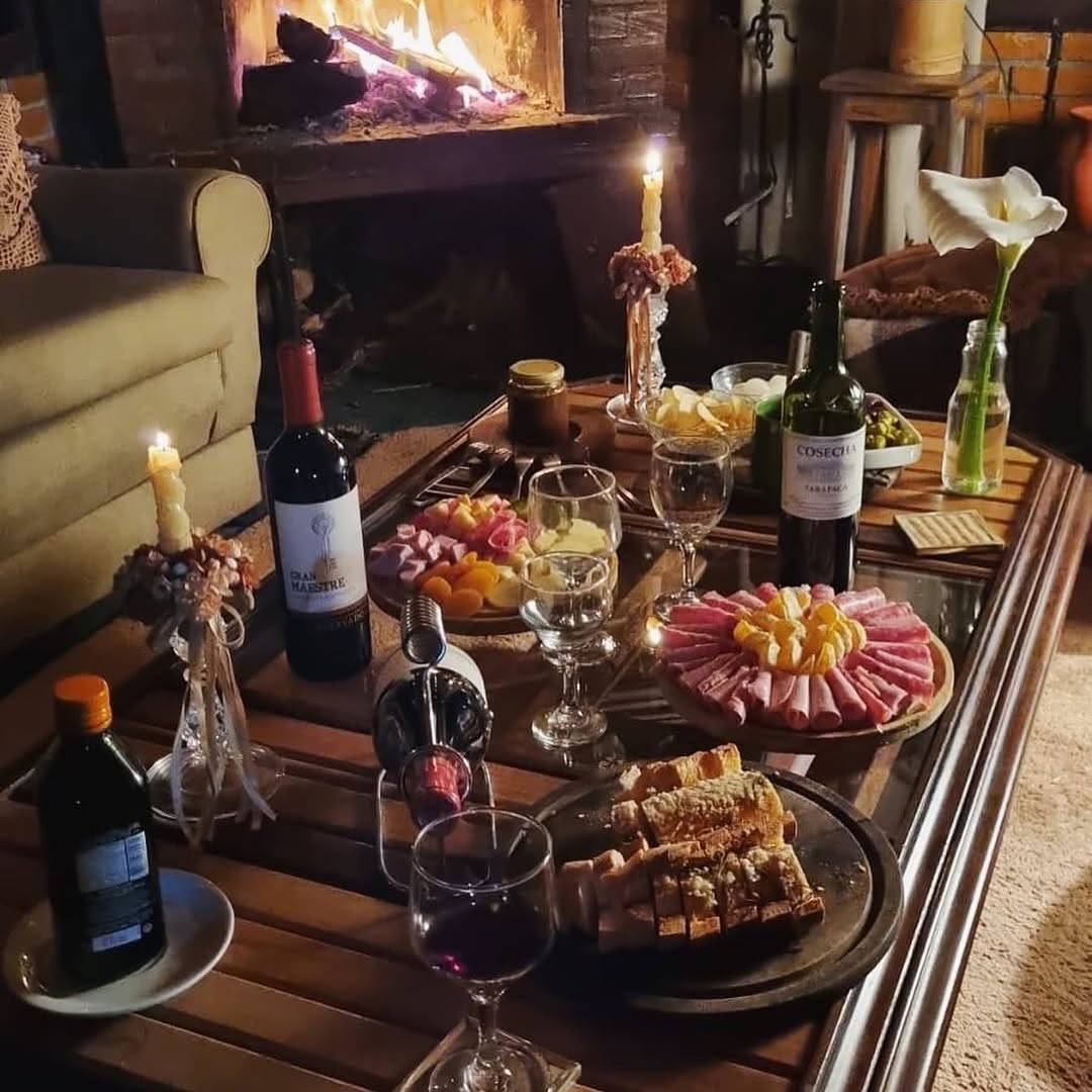 Mesa elegante com tábua de frios artesanais, vinhos selecionados, taças de cristal e velas, em ambiente íntimo com lareira ao fundo