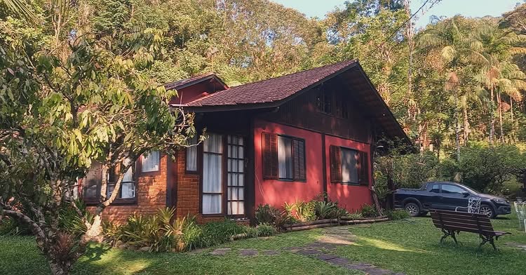 Chalé rústico com fachada em tons terrosos e janelas com venezianas vermelhas, integrado harmoniosamente à paisagem montanhosa