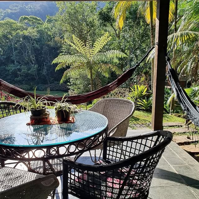 Varanda aconchegante com mesa de vidro, cadeiras de vime e redes, cercada por vegetação tropical luxuriante e palmeiras